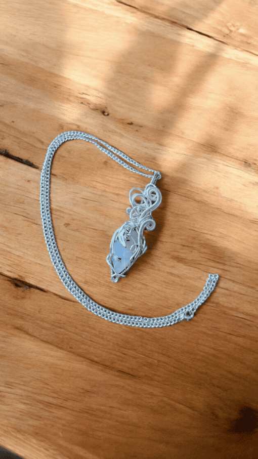 Silver pendant with intricate wire wrapping on a chain, displayed on a wooden surface with sunlight casting shadows.