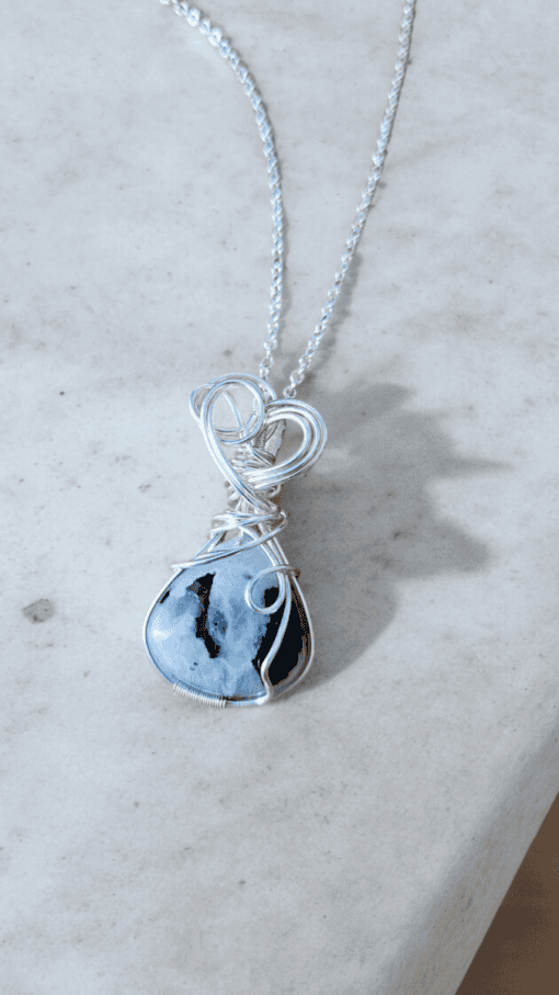 A silver wire-wrapped pendant with a blue and black marbled stone, displayed on a light gray marble surface.