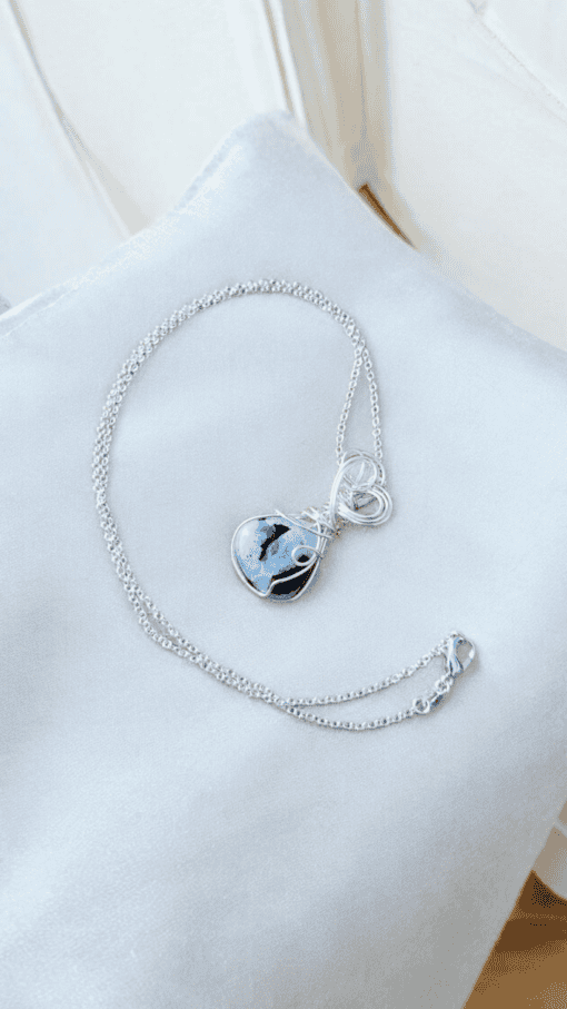 Silver necklace with a blue and black heart-shaped stone pendant, displayed on a light gray fabric background.