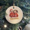 Christmas tree ornament with Santa and Mrs. Claus illustration, hanging among green branches and lit by lights.