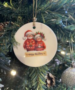 Christmas tree ornament with Santa and Mrs. Claus illustration, hanging among green branches and lit by lights.