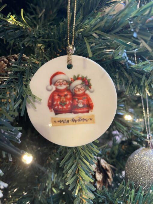 Christmas tree ornament with Santa and Mrs. Claus illustration, hanging among green branches and lit by lights.