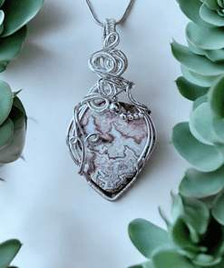 Silver pendant with intricate wirework, featuring a heart-shaped stone with pink and white patterns, surrounded by green leaves.