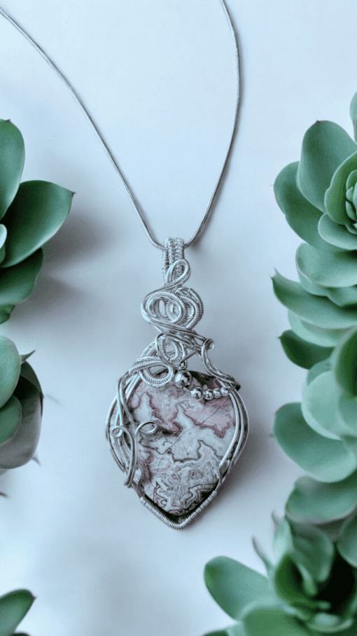 Silver pendant with intricate wirework, featuring a heart-shaped stone with pink and white patterns, surrounded by green leaves.