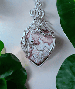 A silver wire-wrapped pendant featuring a marbled stone with intricate patterns, set against a backdrop of green leaves.