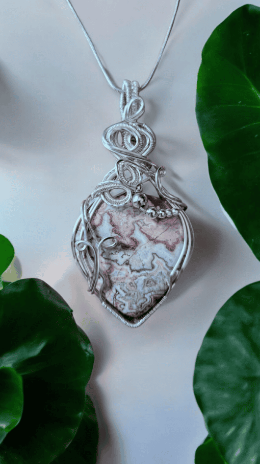 A silver wire-wrapped pendant featuring a marbled stone with intricate patterns, set against a backdrop of green leaves.