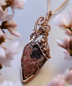 A pendant with intricate wire wrapping holds a dark pink and black gemstone, surrounded by soft, blooming flowers.