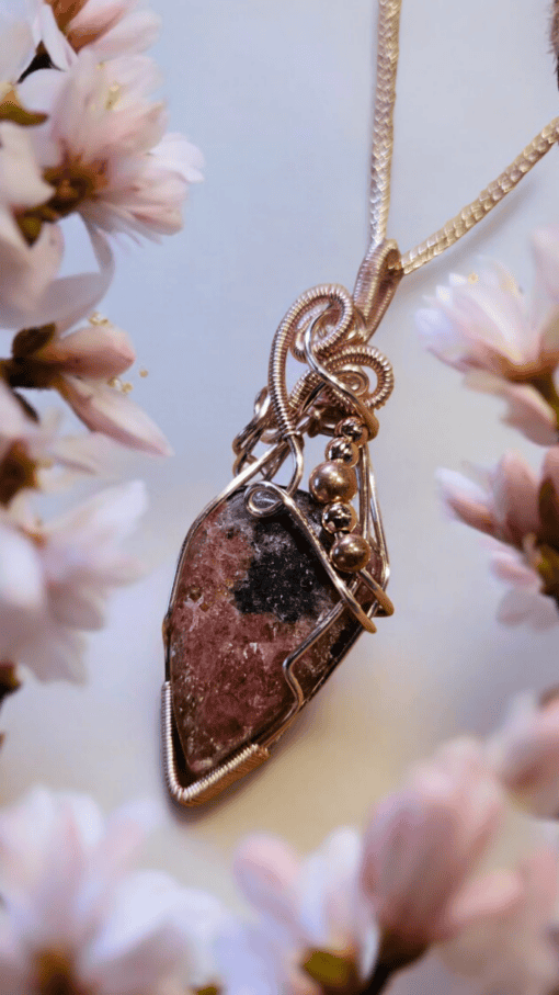 A pendant with intricate wire wrapping holds a dark pink and black gemstone, surrounded by soft, blooming flowers.