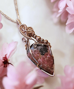 A wire-wrapped stone pendant with intricate design surrounded by pink flowers on a light background.