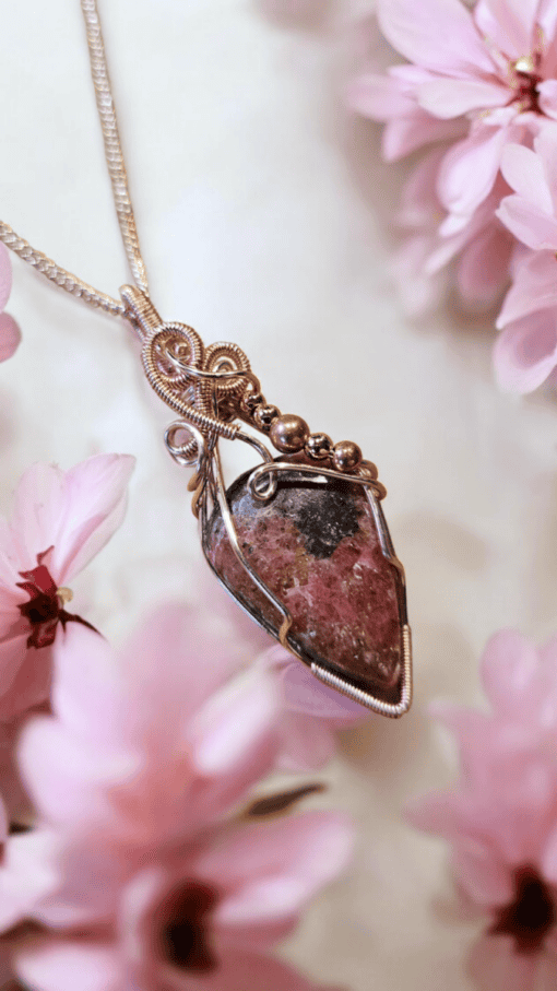 A wire-wrapped stone pendant with intricate design surrounded by pink flowers on a light background.