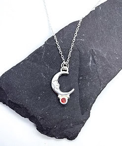 Silver necklace with moon pendant and red gemstone, displayed on a black slate background.