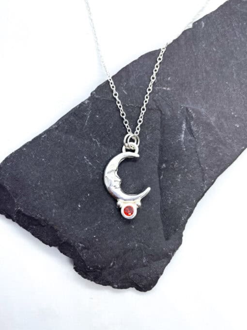 Silver necklace with moon pendant and red gemstone, displayed on a black slate background.