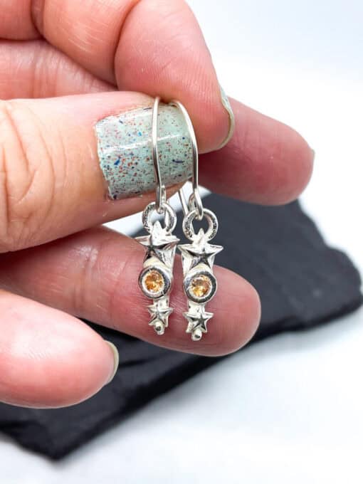 Hand holding star-shaped silver earrings with amber gemstones against a blurred background.