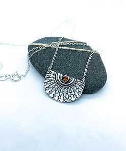 Silver pendant necklace with intricate sunburst design and amber gem centerpiece, displayed on a smooth gray stone background.