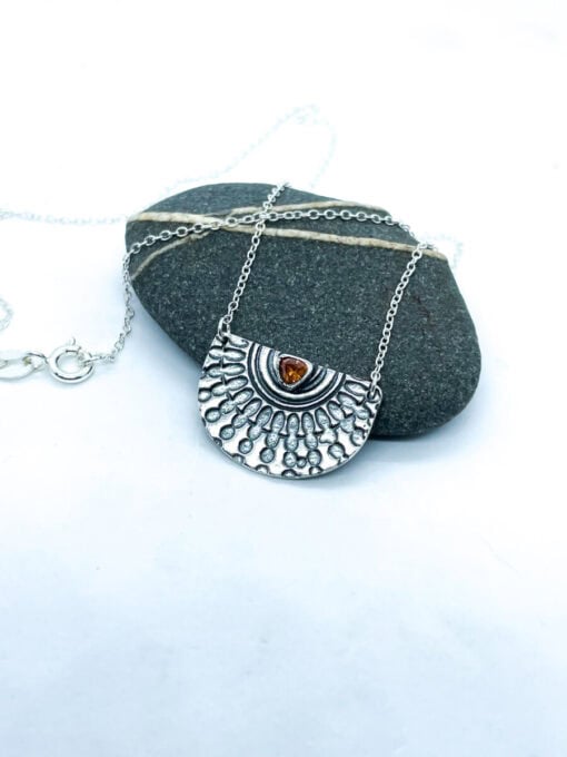 Silver pendant necklace with intricate sunburst design and amber gem centerpiece, displayed on a smooth gray stone background.