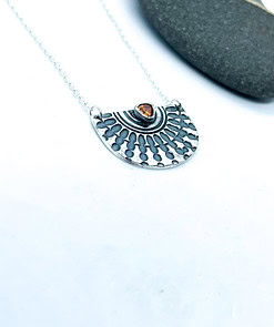 Silver pendant necklace with intricate pattern and amber stone at the center, placed on a white background near a black stone.