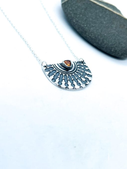 Silver pendant necklace with intricate pattern and amber stone at the center, placed on a white background near a black stone.