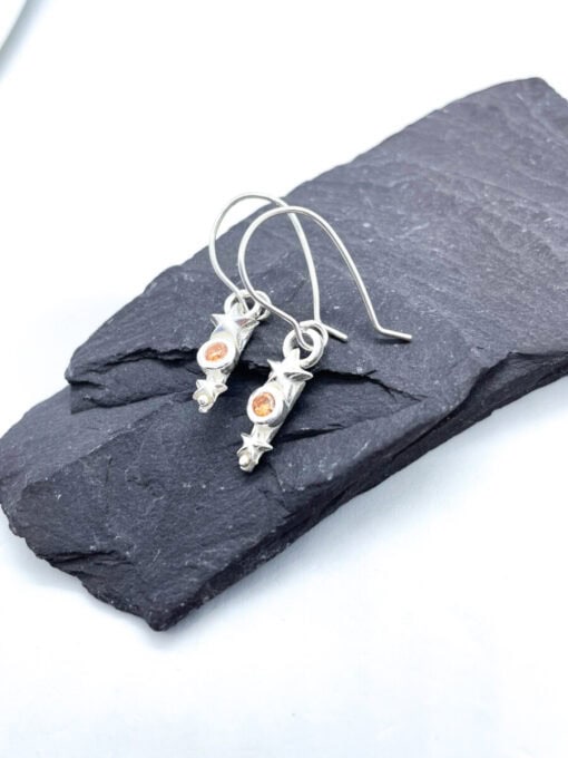 Silver star and scroll earrings with small orange gemstones, displayed on a dark slate platform against a light background.