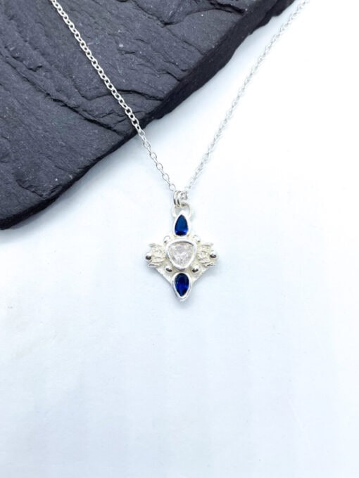 Silver necklace with blue and clear gemstone pendant lying on a dark surface.