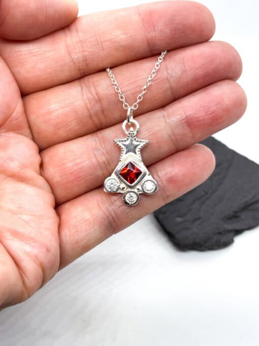 Hand holding a silver star pendant with a red gem and three smaller white gems on a silver chain.