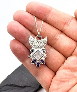 A hand holding a silver pendant necklace with intricate designs and gemstones, including a central square stone and teardrop-shaped amethysts.