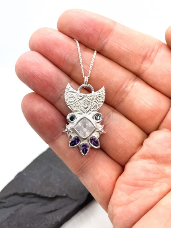 A hand holding a silver pendant necklace with intricate designs and gemstones, including a central square stone and teardrop-shaped amethysts.