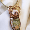 Copper wire-wrapped pendant with a green stone and white flower detailing, set against a white fabric background.