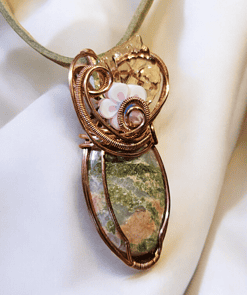 Copper wire-wrapped pendant with a green stone and white flower detailing, set against a white fabric background.