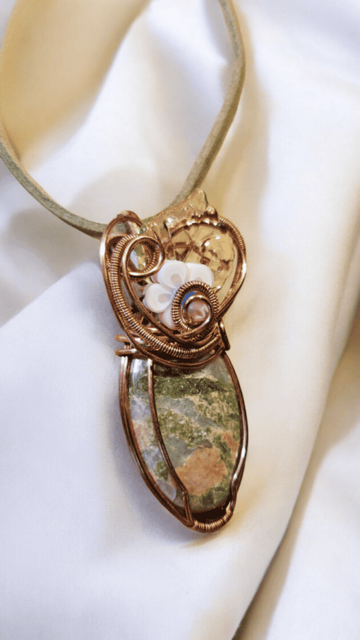 Copper wire-wrapped pendant with a green stone and white flower detailing, set against a white fabric background.