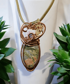 Wire-wrapped pendant with green and brown stone, adorned with intricate designs, displayed between two green plants.