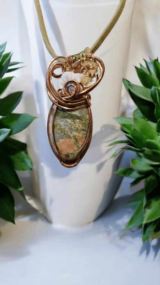 Wire-wrapped pendant with green and brown stone, adorned with intricate designs, displayed between two green plants.