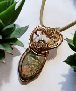 Green and brown stone pendant wrapped in intricate gold wire, adorned with a white flower, placed between two succulent plants.