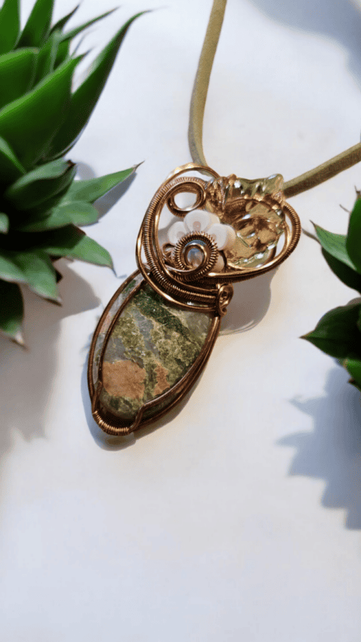 Green and brown stone pendant wrapped in intricate gold wire, adorned with a white flower, placed between two succulent plants.