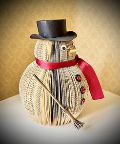 Snowman decoration made from book pages, with a black top hat, red scarf, buttons, and a broom, on a white surface.