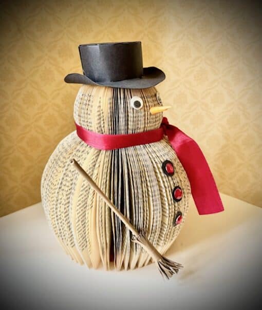 Snowman decoration made from book pages, with a black top hat, red scarf, buttons, and a broom, on a white surface.