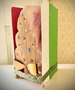 A folded book sculpture resembling a Christmas tree adorned with colorful star ornaments and a gold ribbon, encased in green covers.