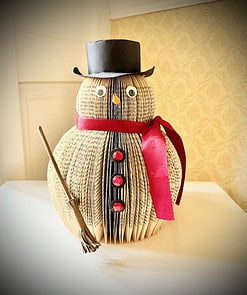 Snowman crafted from folded book, adorned with a red ribbon, buttons, black top hat, and holding a small broom.