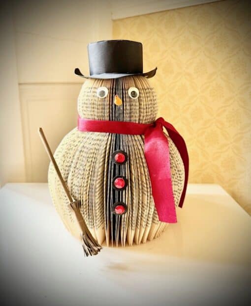 Snowman crafted from folded book, adorned with a red ribbon, buttons, black top hat, and holding a small broom.