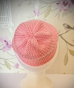 Pink and white knitted beanie displayed on a mannequin head against a floral wallpaper background.