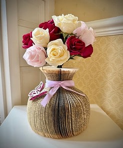 Bouquet of red, white, and pink roses in a vase made from a folded book, adorned with a lavender ribbon and a butterfly decoration.