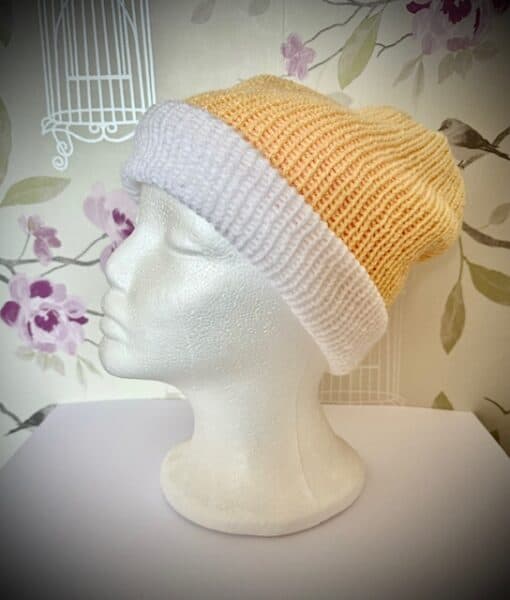 A mannequin head with a knitted beanie featuring a yellow top and white folded brim, set against a floral wallpaper background.