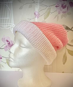 White mannequin head wearing a pink and white gradient knitted beanie, in front of floral wallpaper with birdcage design.