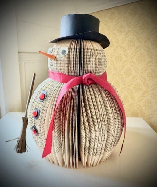 Snowman decoration made from folded book pages, adorned with a black hat, red scarf, button eyes, and a broomstick.