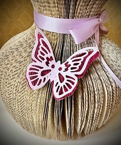 Book pages folded into a dress shape, adorned with a pink butterfly cutout and tied with a pink ribbon.