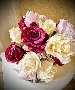 Bouquet of pink, white, and purple crepe paper roses in a white vase, displayed against a patterned wallpaper background.