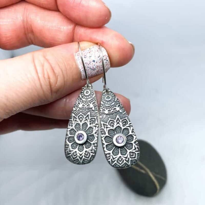 Hand holding silver drop earrings with intricate floral patterns and a small central gem over a blurred background.