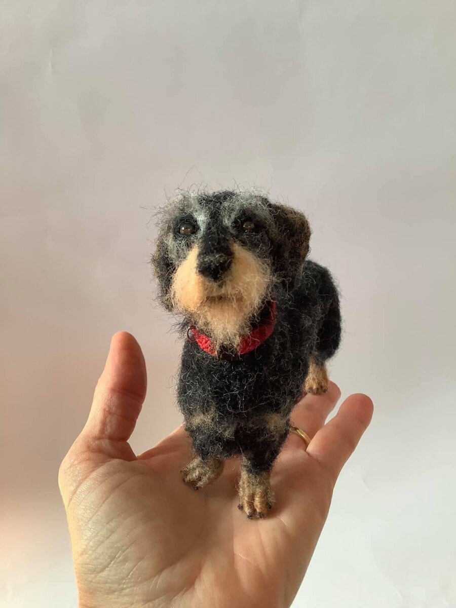 A small felted dog sculpture with a black and brown coat and a red collar is held in a person's hand.