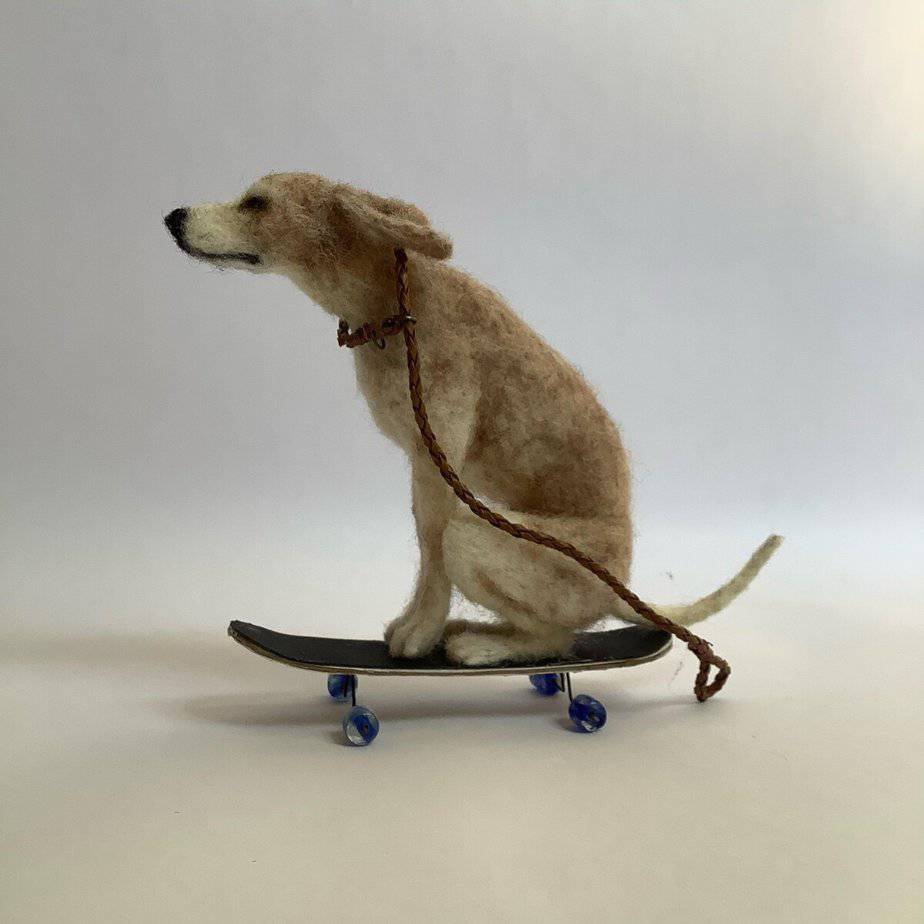 Felt sculpture of a dog sitting on a skateboard with a rope around its neck, against a plain background.