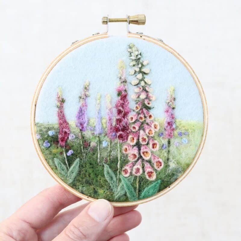 Hand holding an embroidery hoop featuring a felted wool design of blooming pink and purple flowers with green leaves and stems.