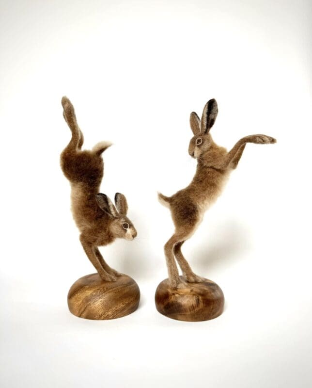 Two felted rabbit sculptures, each balanced on wooden bases, with one standing on one leg and the other in a handstand pose.